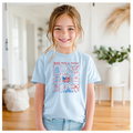 Young girl wearing a light blue t-shirt with a graphic design, standing in a room with wooden flooring and white walls.